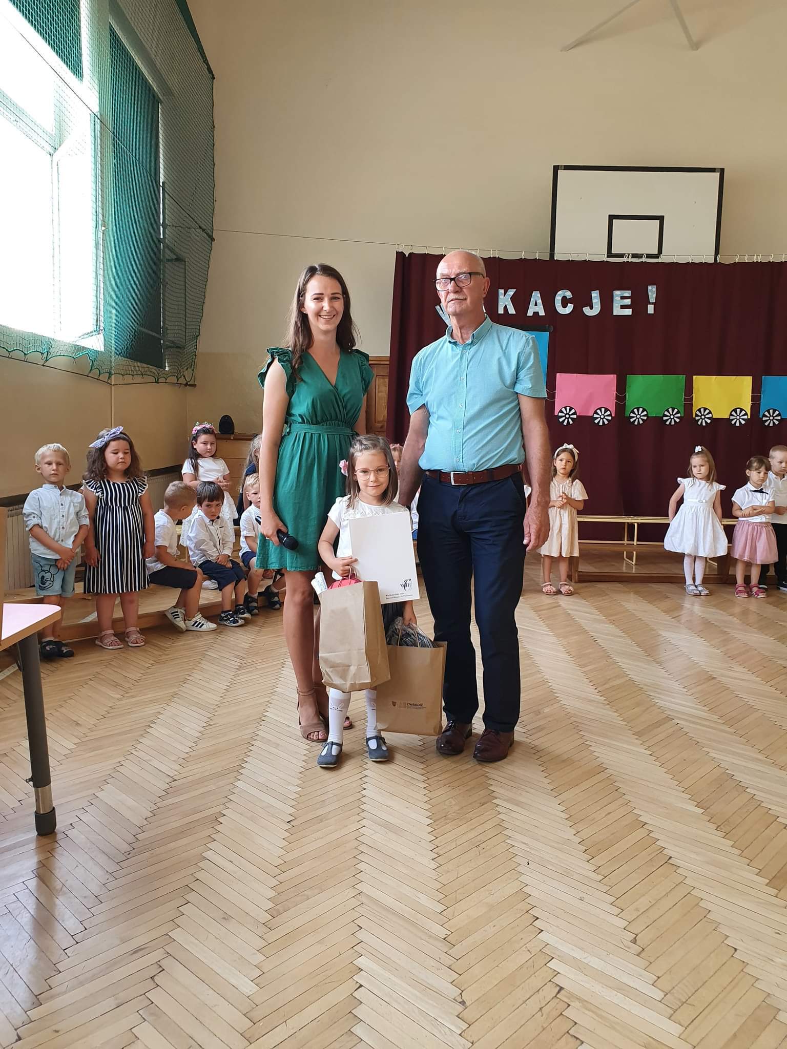 Na zdjęciu widać małą dziewczynkę (w wieku przedszkolnym), jej przedszkolną opiekunkę oraz dyrektora CWRKDiZ w Lesznie.
Dziewczynka trzyma w ręce nagrody w papierowych torbach.