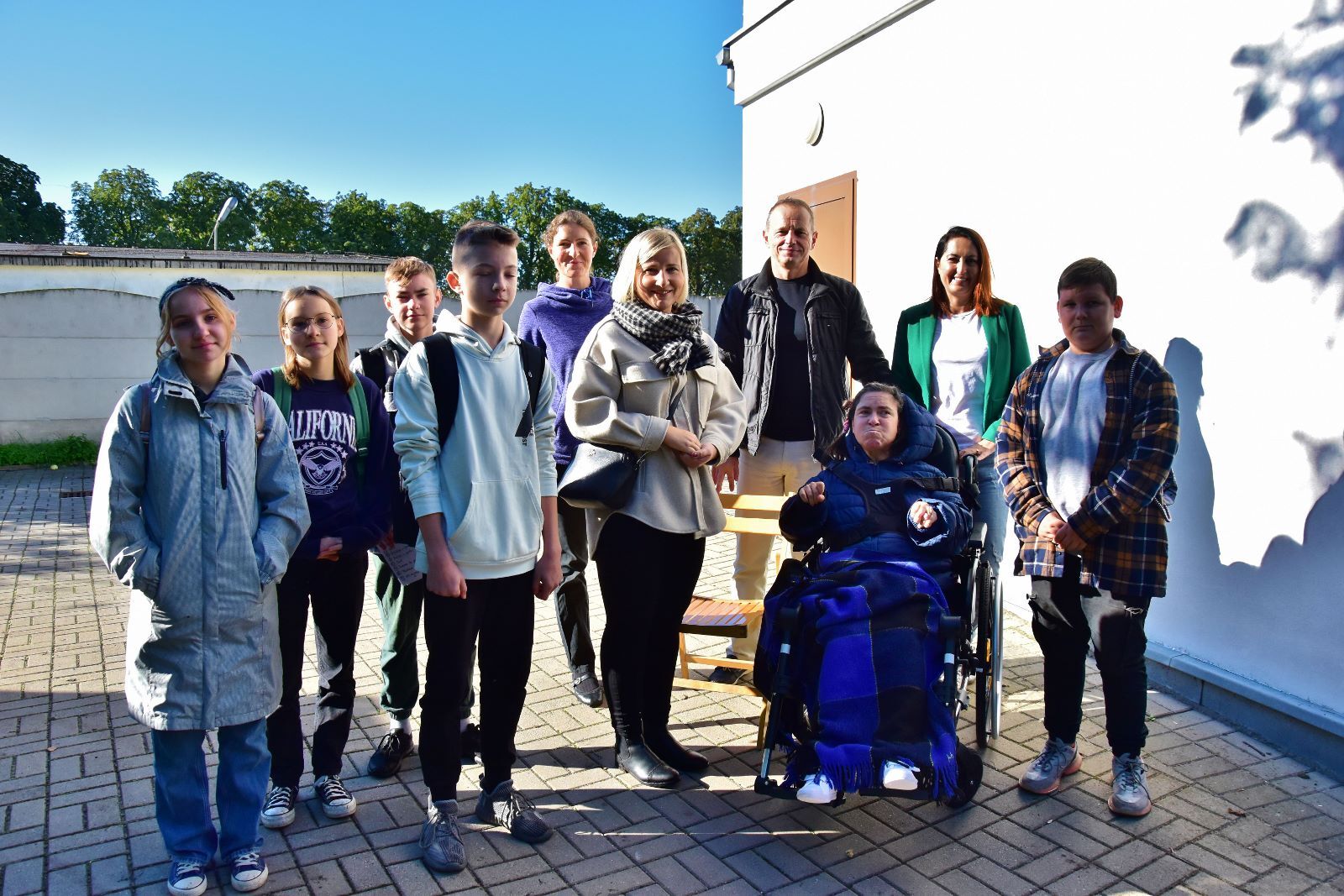 Na zdjęciu widać grupę młodych osób (uczniów szkół podstawowych) wraz z opiekunami oraz osobą na wózku inwalidzkim.