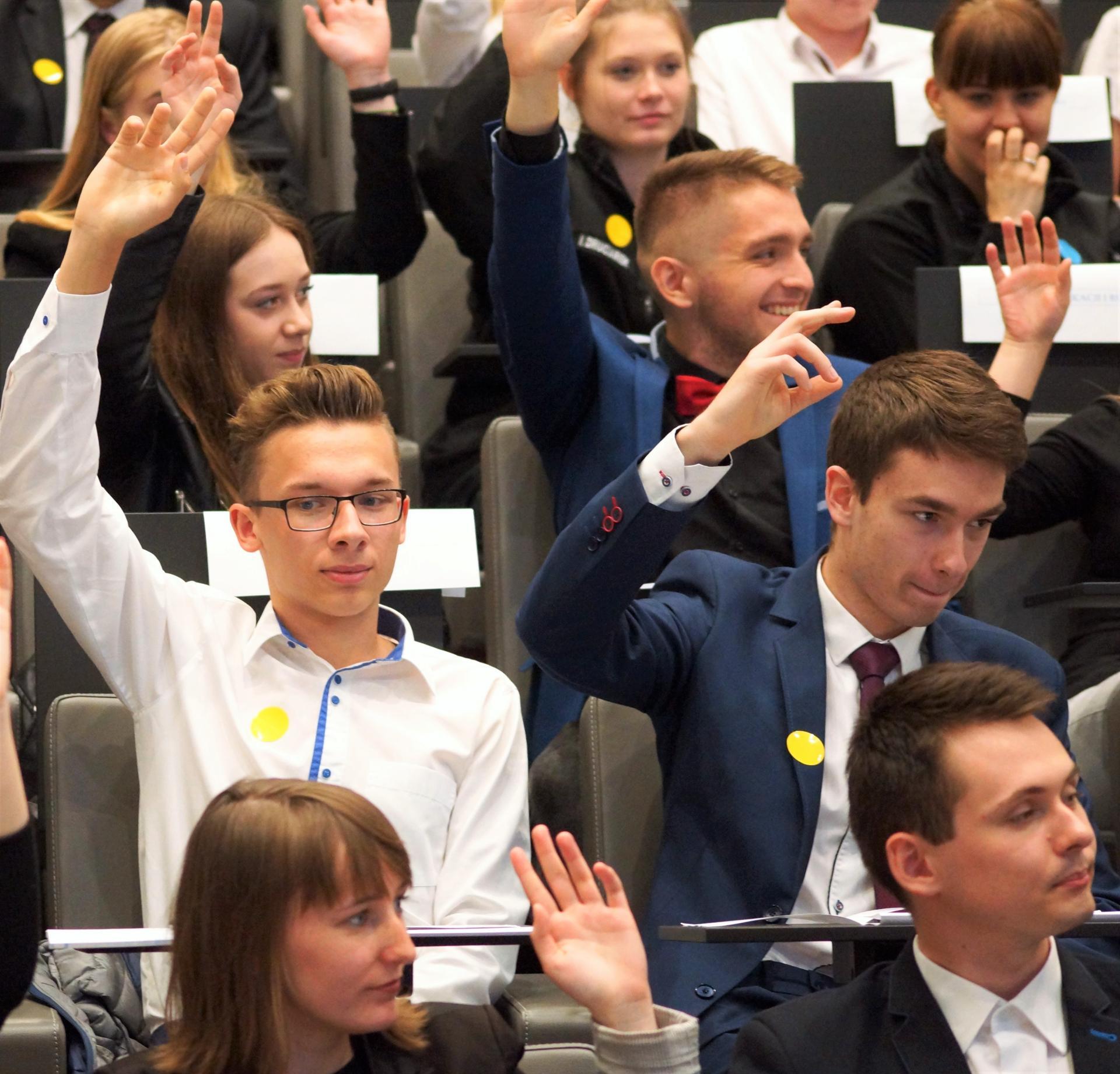 Na fotografii widać młodych ludzi siedzących na sali posiedzeń i podnoszących rękę do góry - najprawdopodobniej biorą udział w głosowaniu.