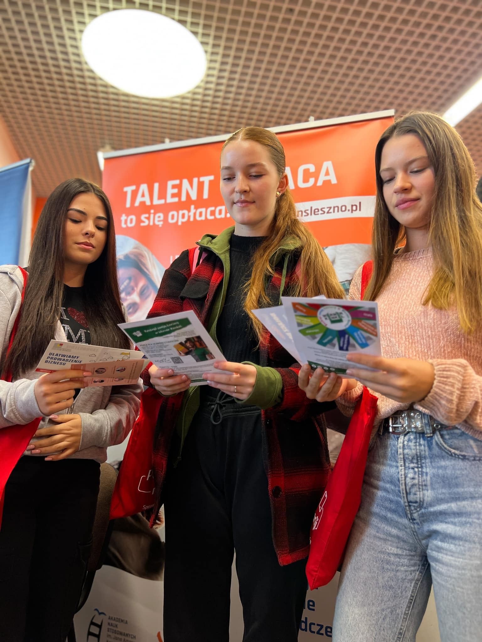 Trzy młode dziewczyny trzymają w rękach broszury. Za nimi rollup "Talent i praca - to się opłaca!".