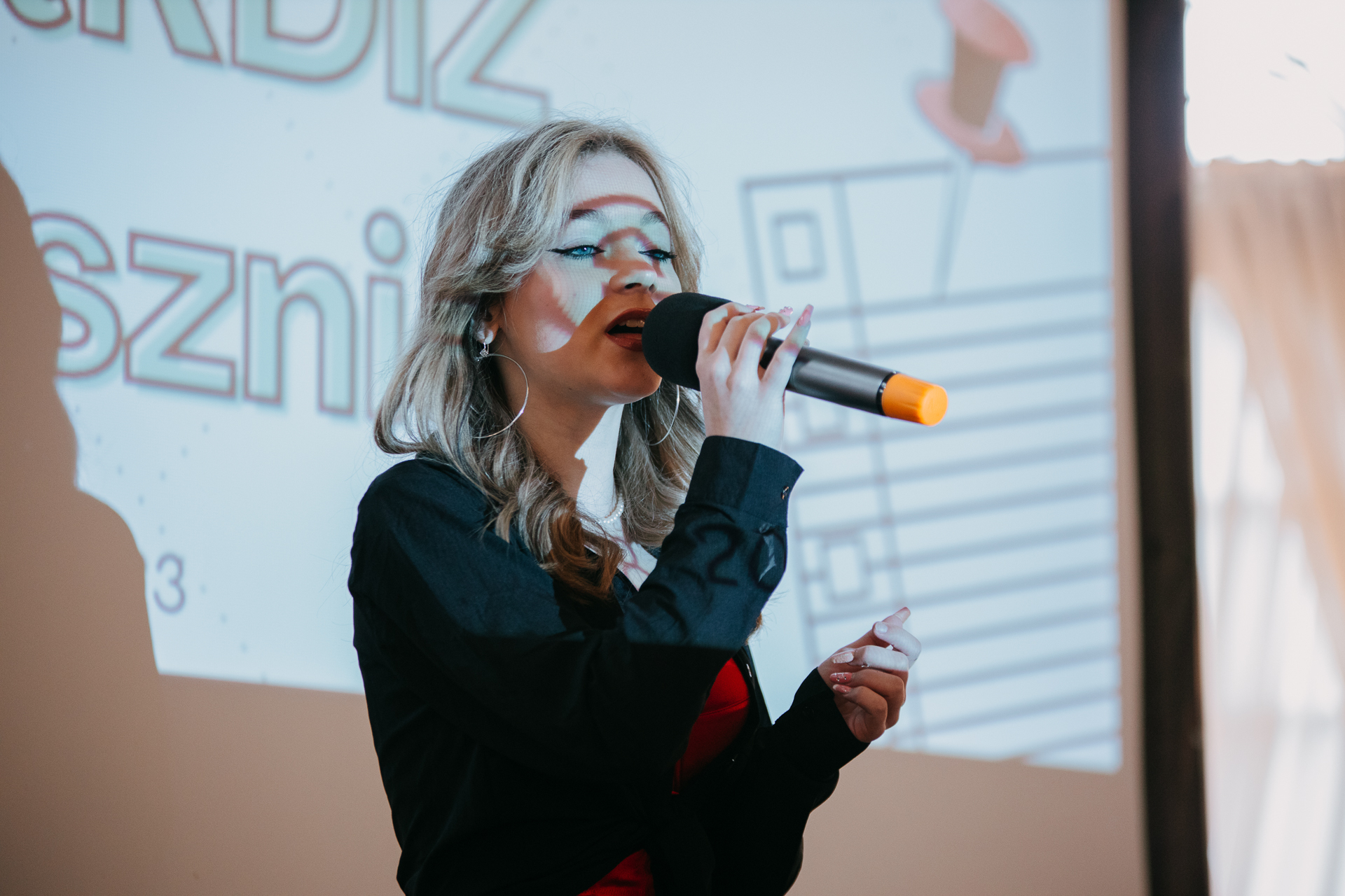Na zdjęciu młoda dziewczyna - Emilia Nowak - stojąca na scenie i śpiewająca do mikrofonu.