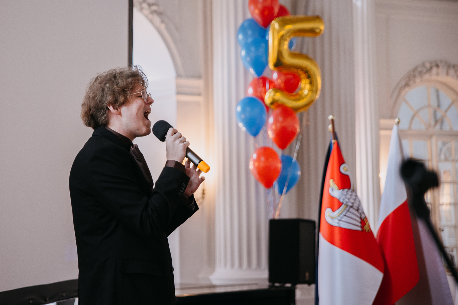 Na scenie młody chłopak, artysta, piosenkarz - Norbert Wronka - ubrany w czarny garnitur śpiewa do mikrofonu.