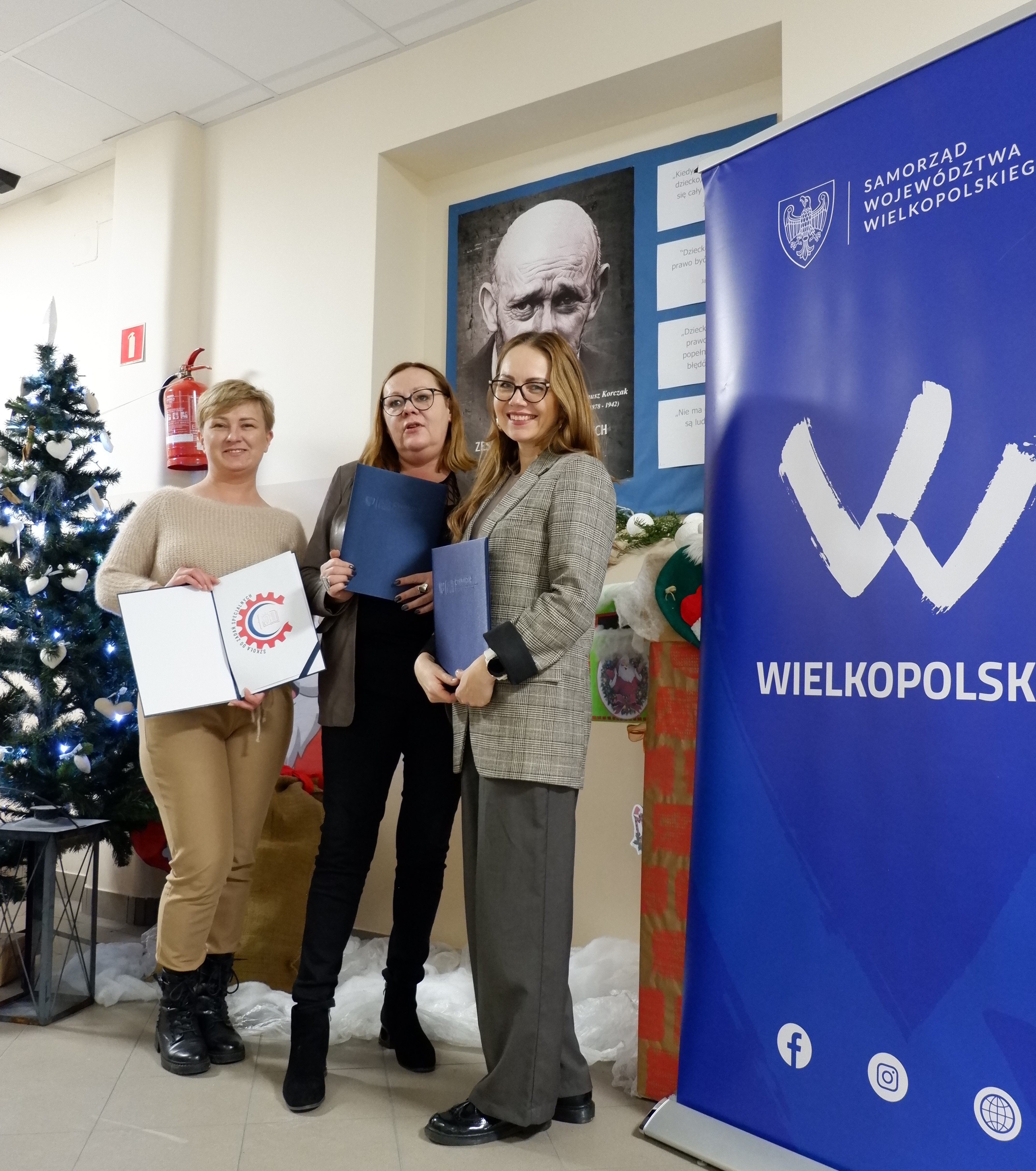 Na zdjęciu widać trzy uśmiechnięte kobiety, które trzymają w rękach podpisane porozumienie oraz zaprojektowany logotyp projektu. W tle rollup Wielkopolski oraz Jan Korczak - patron szkoły.