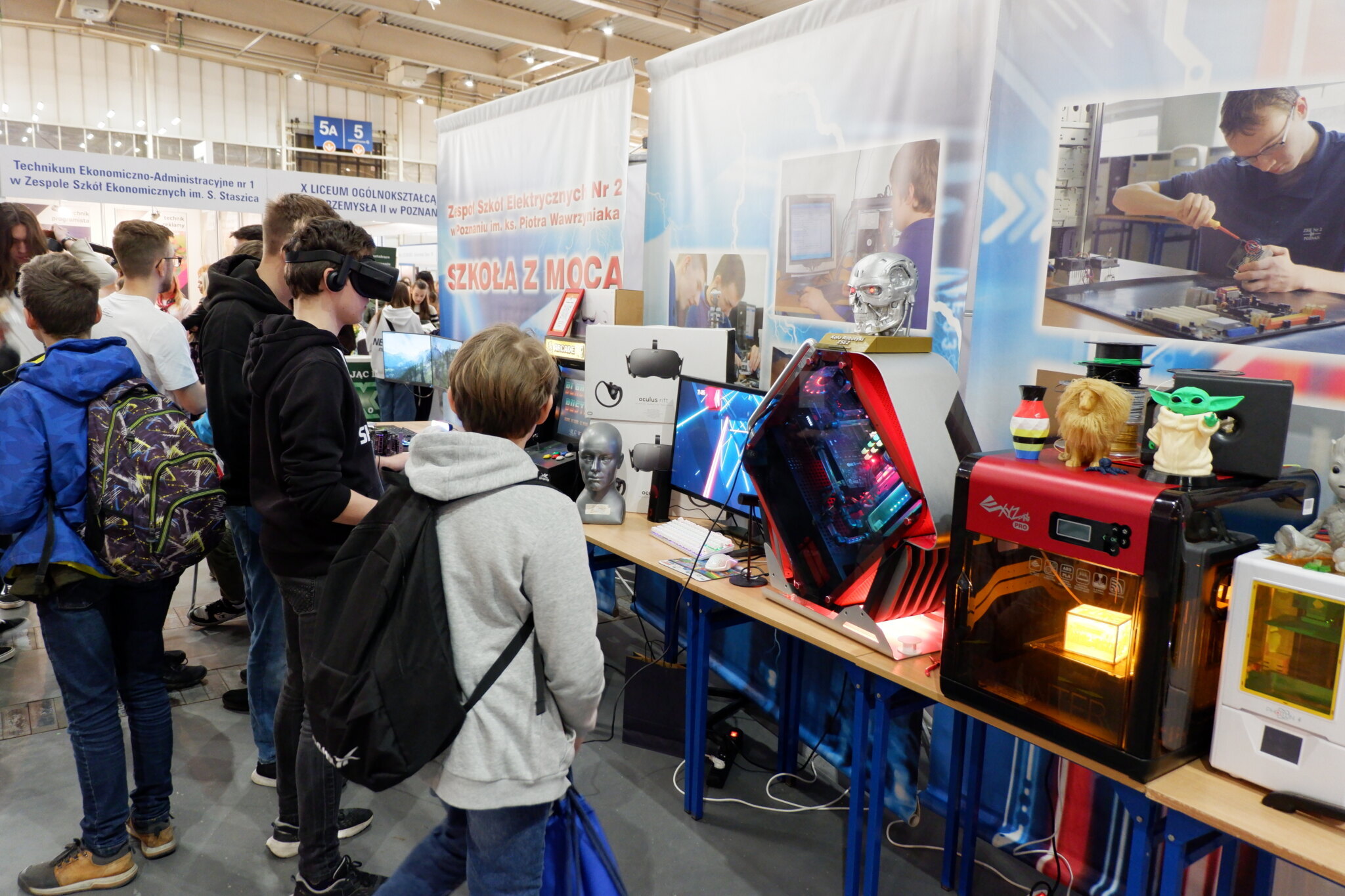 Na zdjęciu widać młodzież stojącą na stoisku interaktywnym podczas Targów Edukacyjnych 2024 w Poznaniu. Niektóre osoby mają założone okulary VR i grają w coś na komputerze.